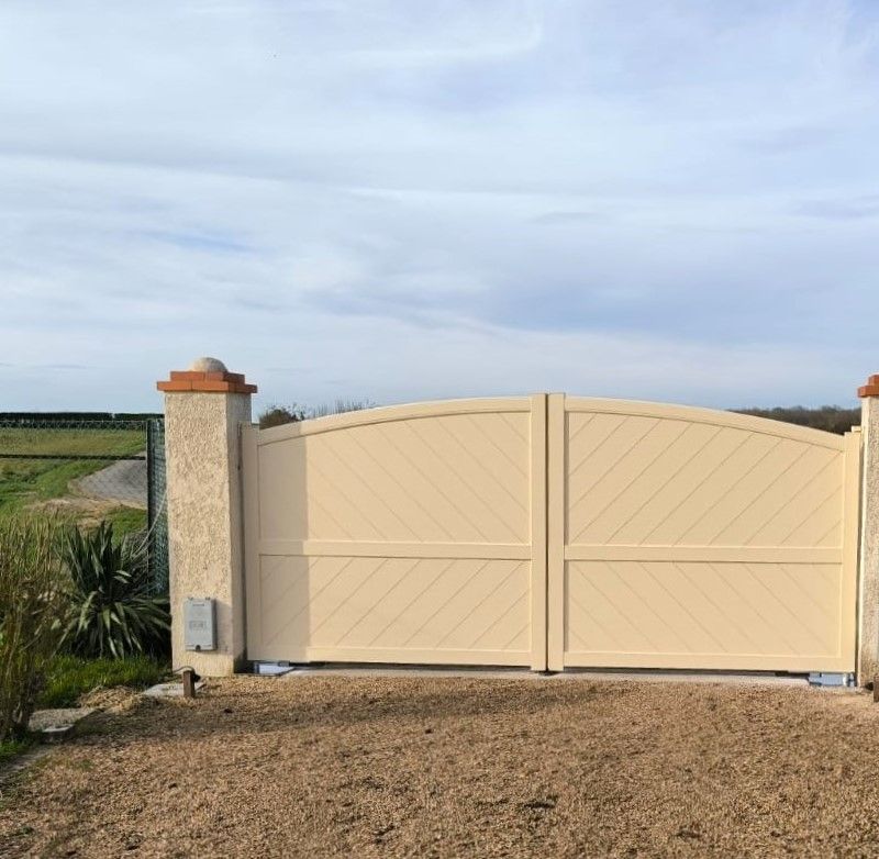 Portail en métal beige avec deux piliers, allée et haie.