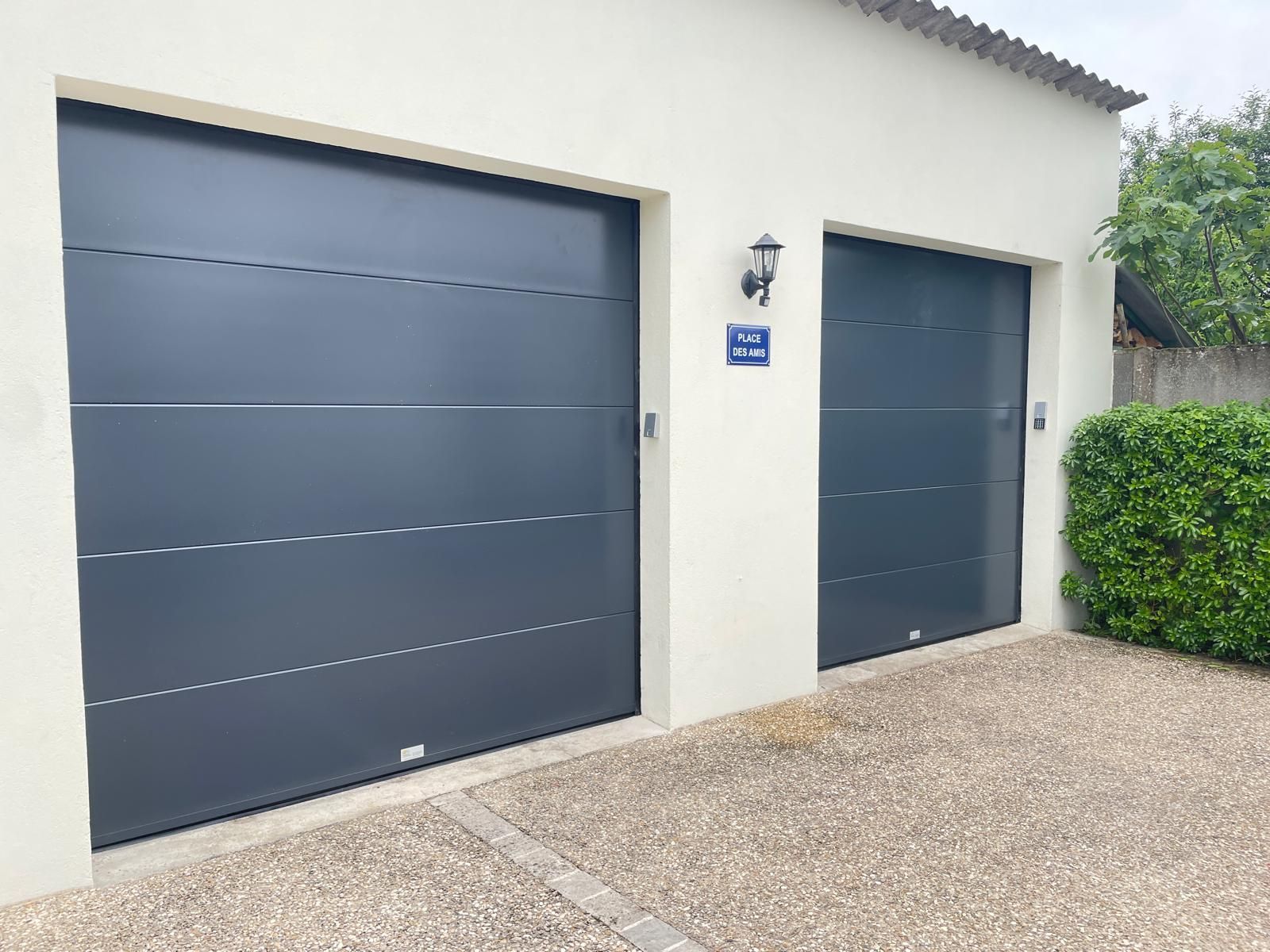 Deux portes de garage gris foncé sur une maison beige clair avec une allée en gravier.