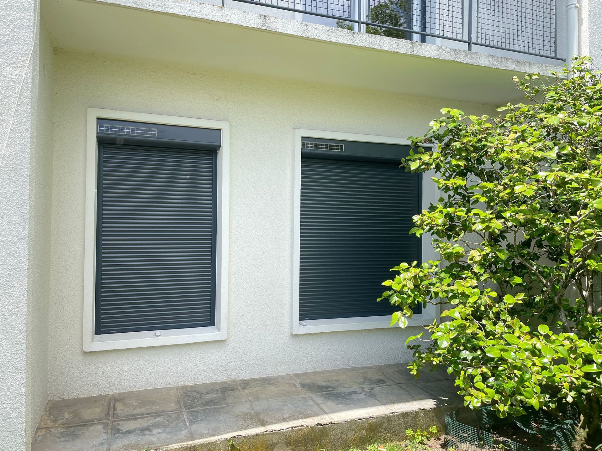 Portes-fenêtres et volets blancs sur un bâtiment en stuc clair, toit en ardoise foncée, lucarne avec volet.