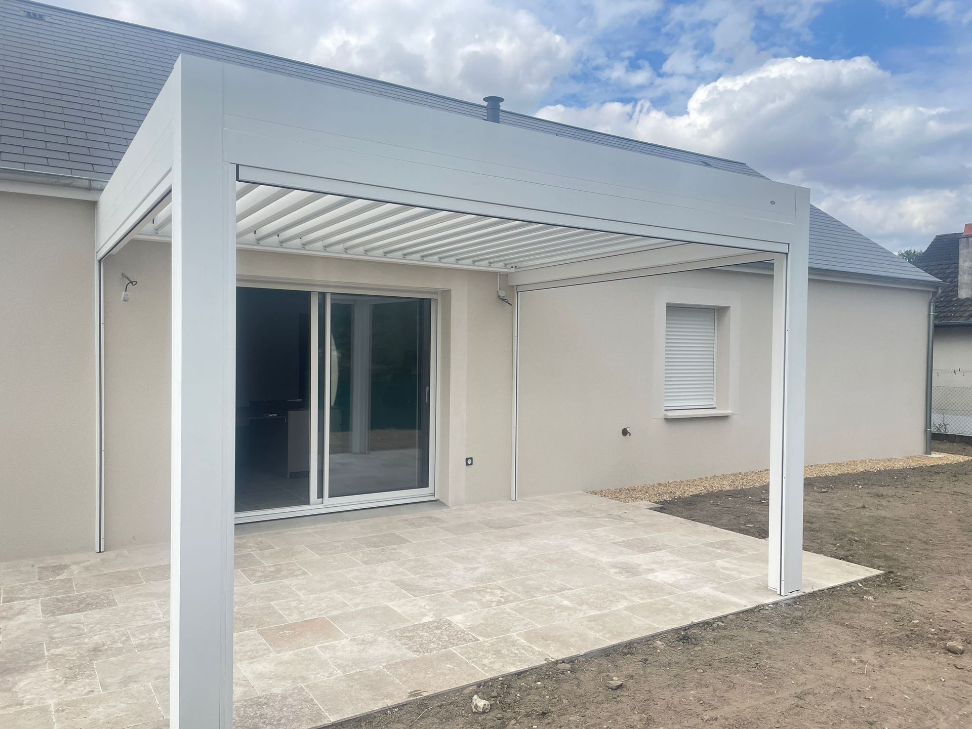 Pergola blanche au-dessus d'une terrasse en pierre, attenante à une maison de couleur claire avec des portes coulissantes en verre.