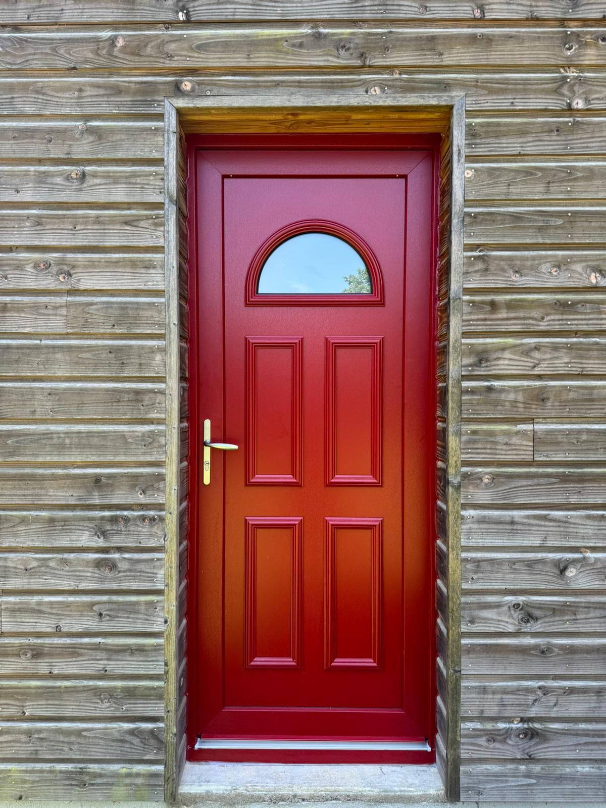 Porte rouge avec fenêtre cintrée, encadrée de bois et adossée à un mur horizontal en planches de bois.