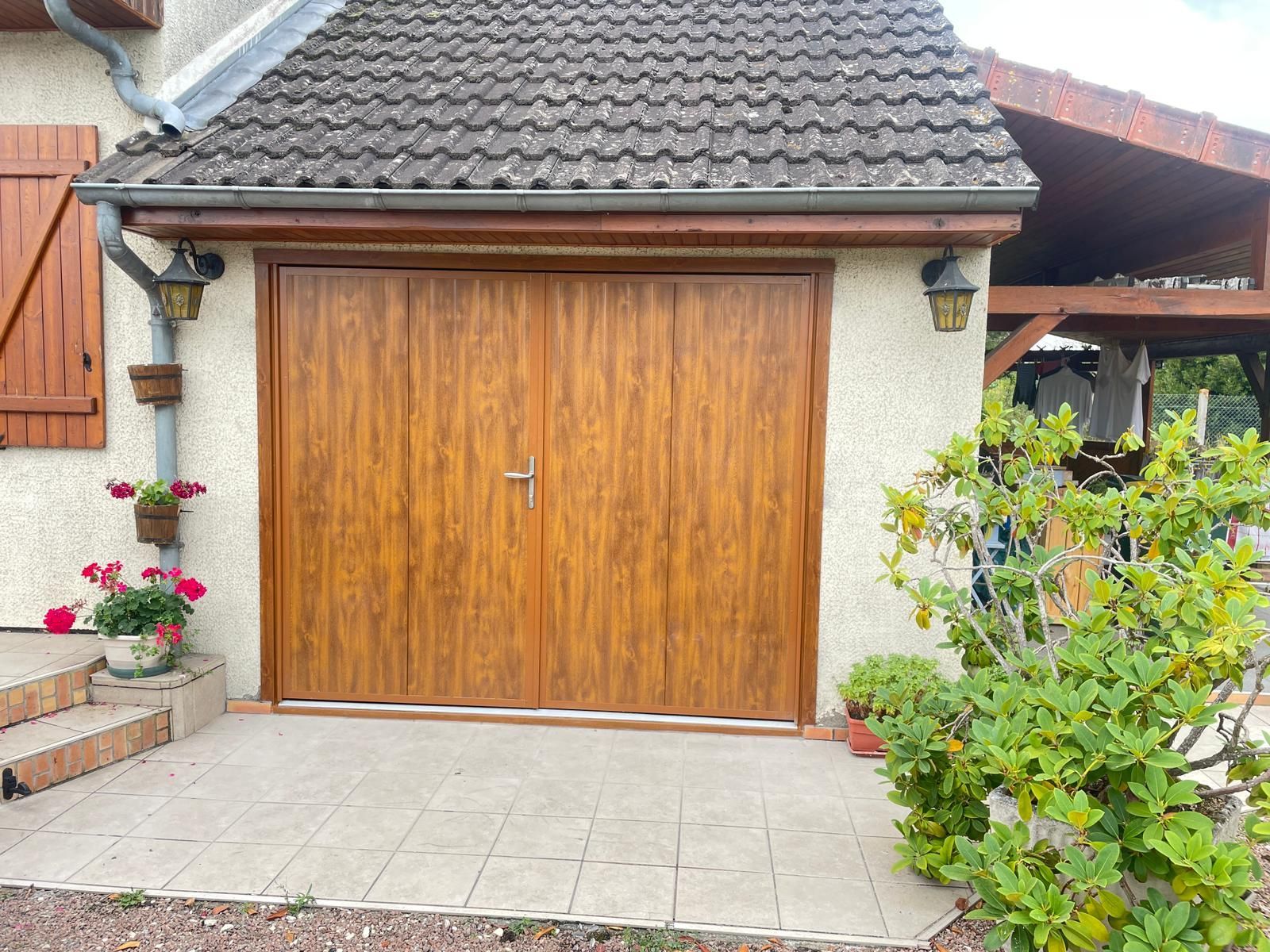 Porte de garage marron sur une maison au toit de tuiles, entourée de plantes et de lumières extérieures.