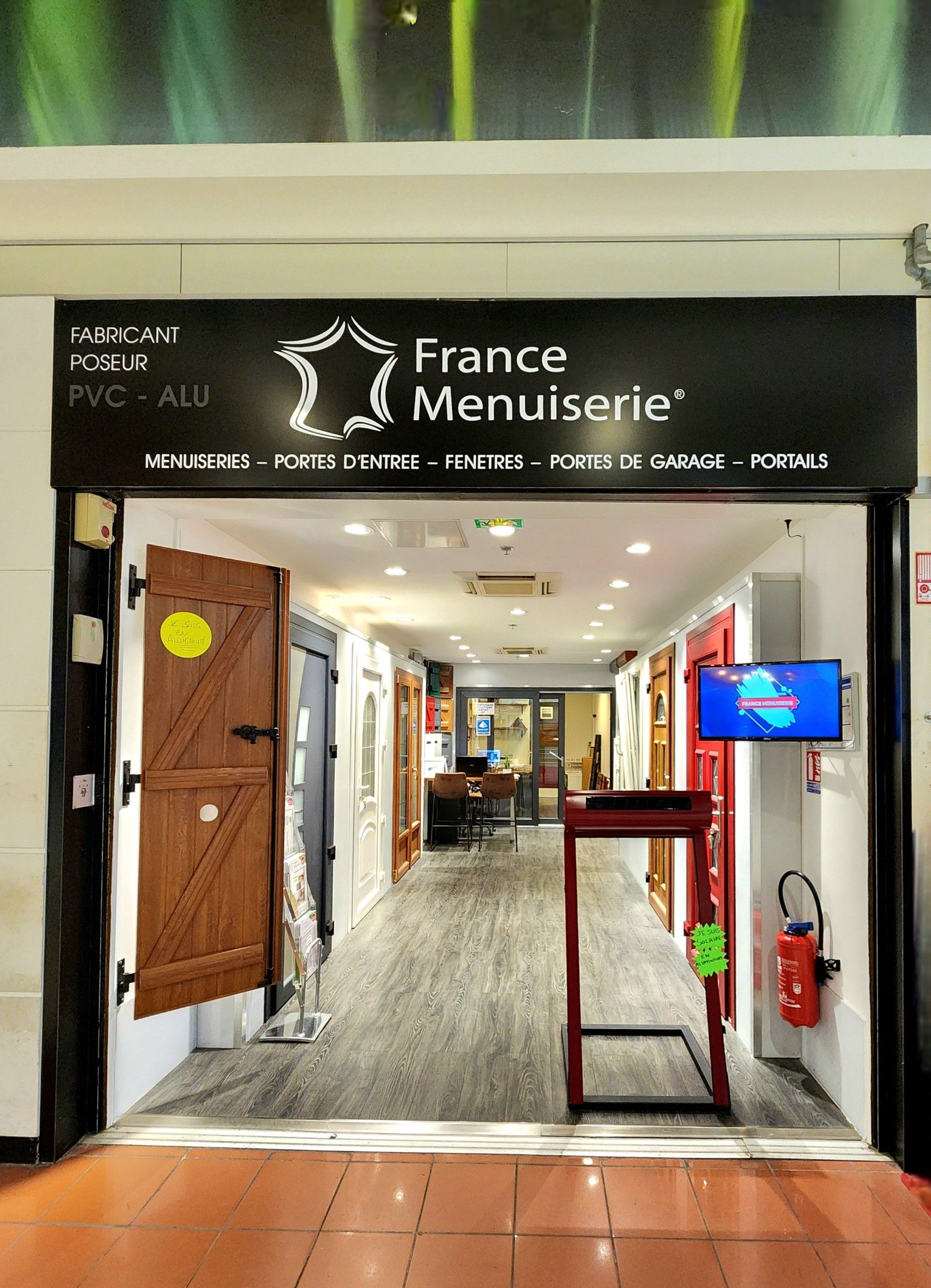 Entrée d'un magasin France Menuiserie, présentant des portes avec des encadrements rouges et marrons, une petite télévision et un extincteur.