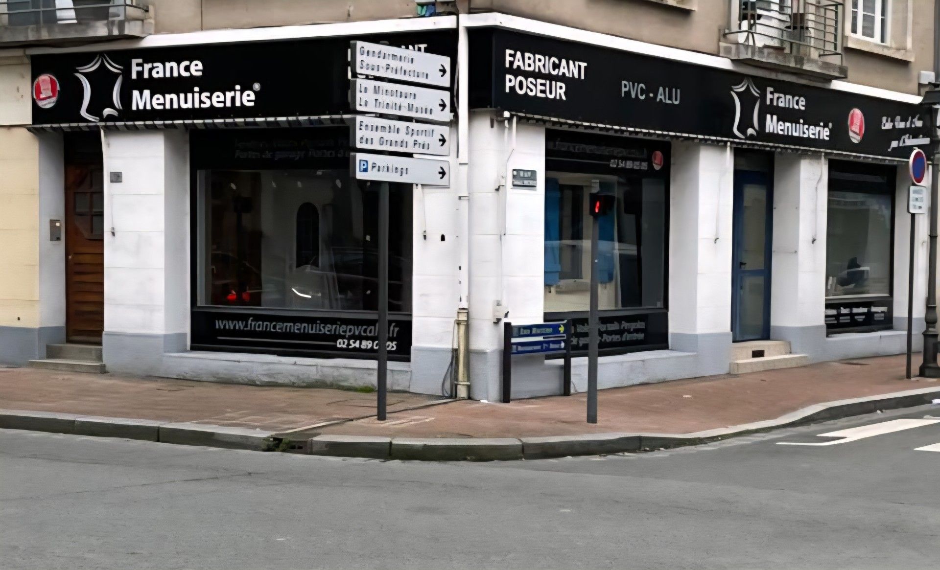 Vitrine d'angle avec l'enseigne « France Menuiserie ». Lettrage foncé sur fond noir, bâtiment blanc et gris.