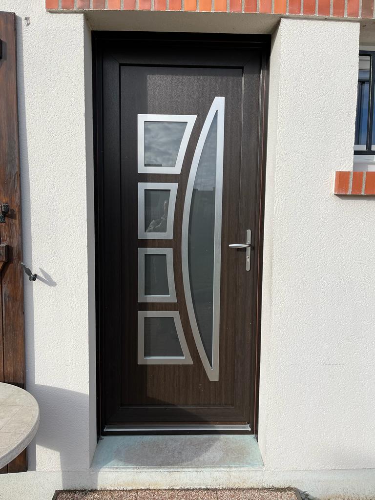 Porte d'entrée brun foncé avec panneaux de verre et garnitures argentées, encastrée dans un mur en stuc blanc.