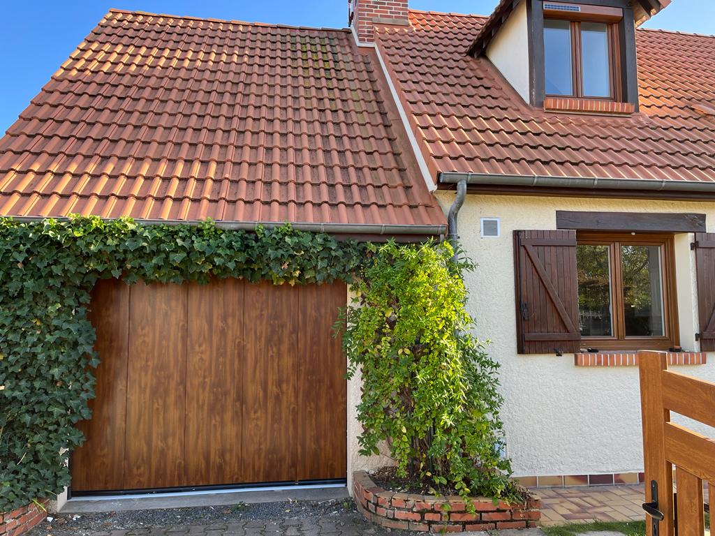 Porte de garage recouverte de lierre sur une maison avec un toit de tuiles et une lucarne.