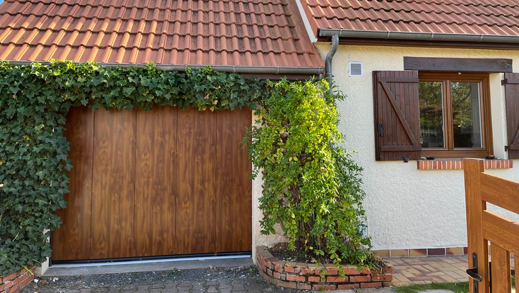 Porte de garage en bois brun avec toit de tuiles rouges recouvert de lierre, et une fenêtre à volets bruns.