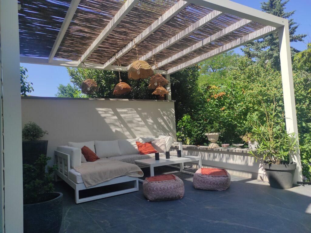Patio avec canapé blanc, table basse et poufs à motifs sous une pergola à claire-voie, entouré de verdure.