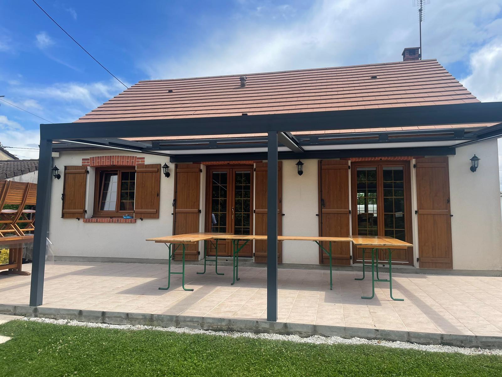Maison blanche aux volets marrons et pergola noire surplombant un patio avec des tables, par une journée ensoleillée.