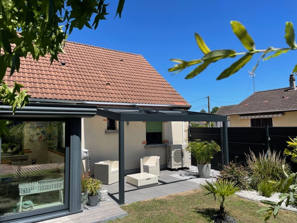 Terrasse extérieure avec pergola grise, mobilier blanc et maison au toit de tuiles rouges sous un ciel bleu azur.