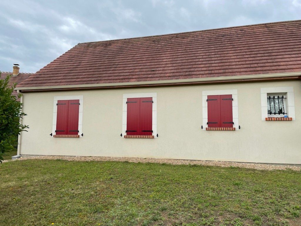 Maison beige avec volets rouges et tuiles au toit, pelouse verte devant, ciel nuageux.