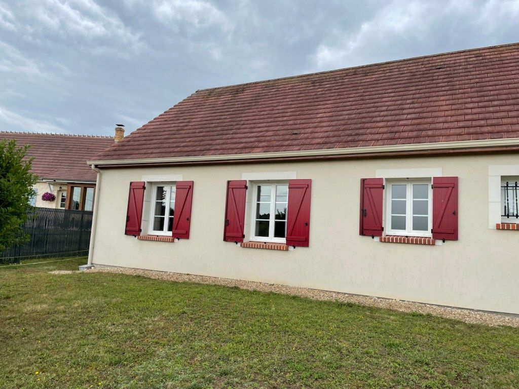 Maison beige aux volets rouges, au toit rouge et aux encadrements de fenêtres blancs, se détachant sur un ciel nuageux et une pelouse verte.