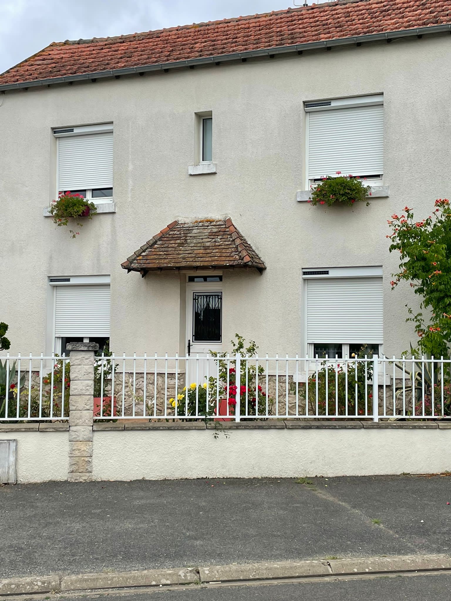 Maison blanche à deux étages avec toit de tuiles rouges, volets blancs, petit porche, clôture blanche et jardinières.