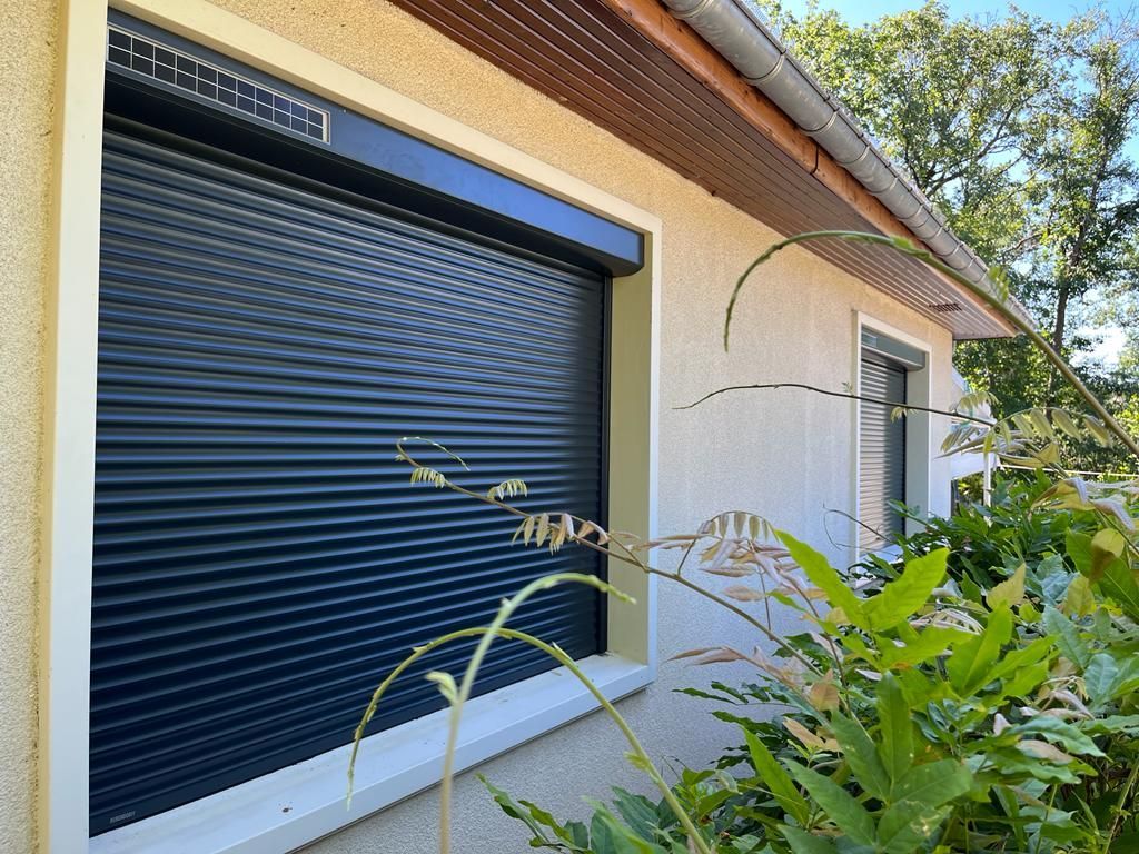 Fenêtre avec volet roulant gris fermé, portes doubles à cadre blanc et volets en bois brun.