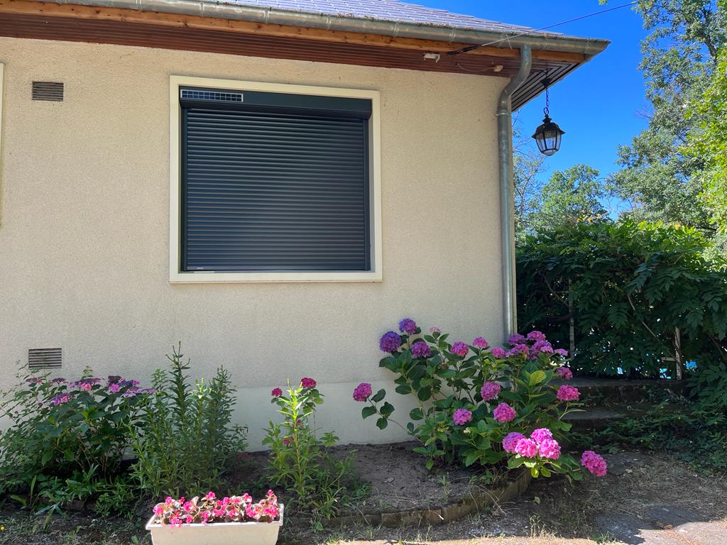Extérieur d'un bâtiment beige avec un volet noir fermé, entouré de fleurs roses et de verdure sous un ciel bleu.