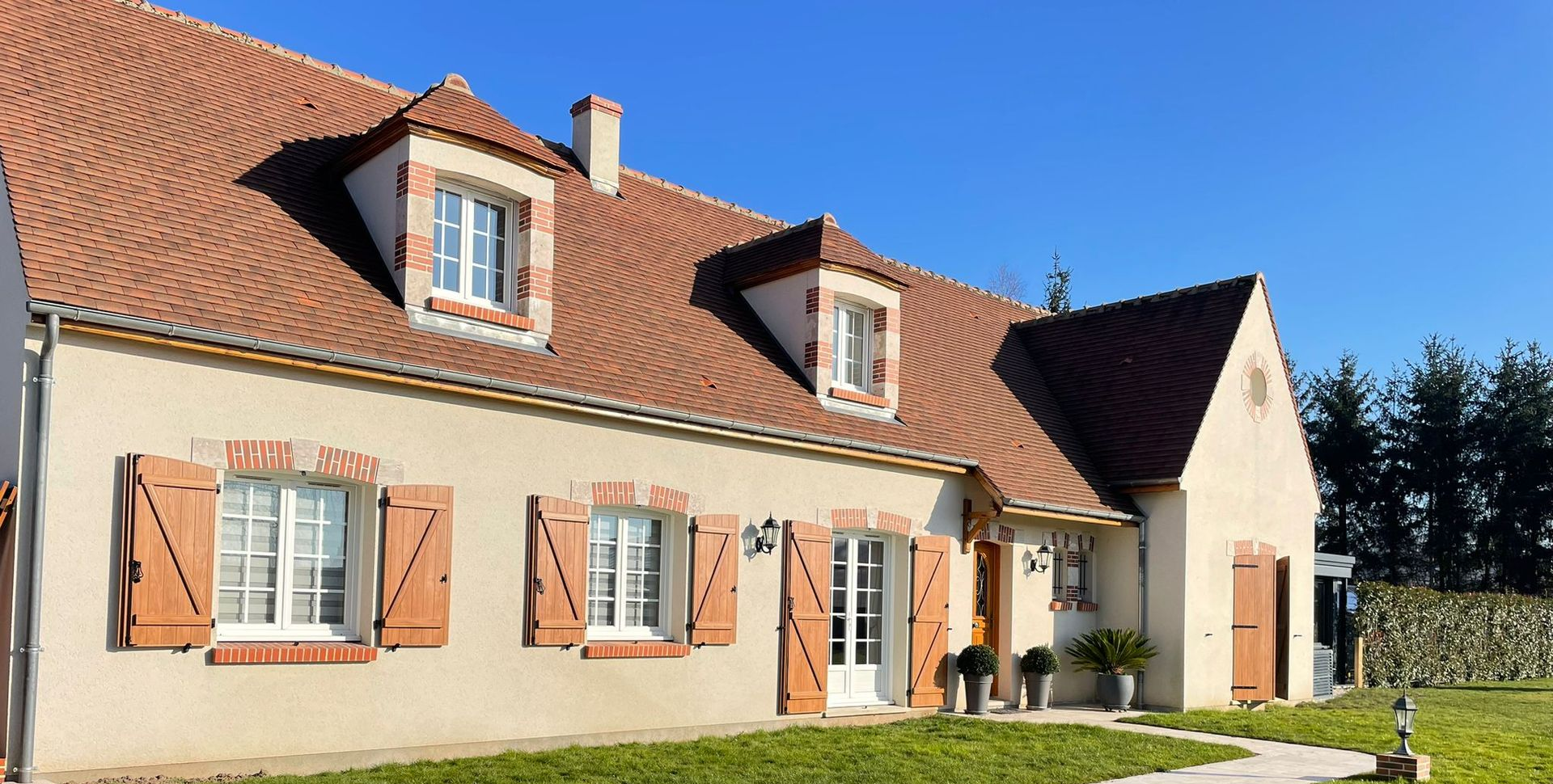 Maison au toit de tuiles rouges, murs beiges, volets et fenêtres marron. Ciel d'un bleu éclatant.