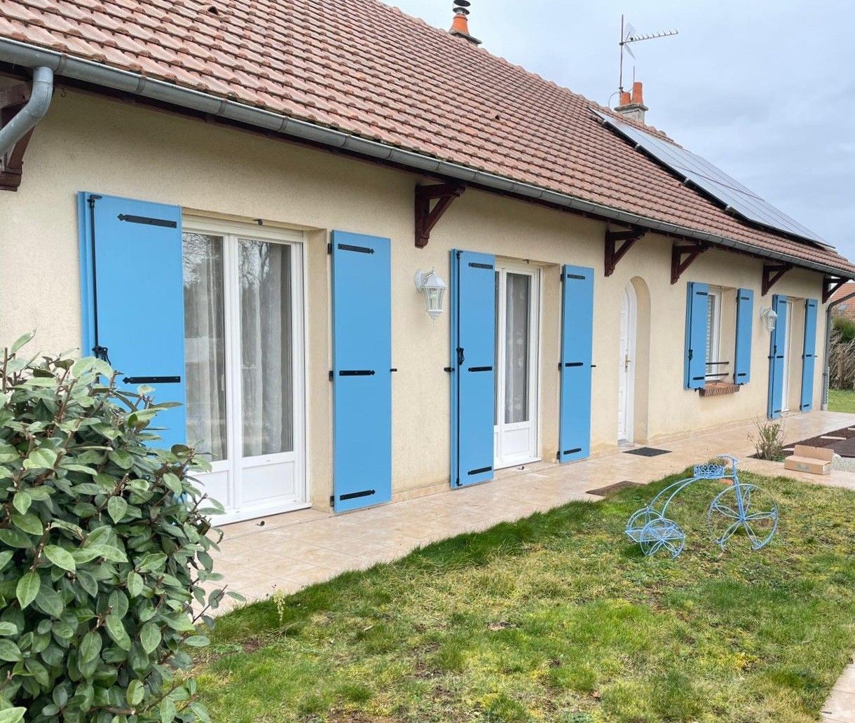 Deux fenêtres à volets rouges sur une maison en stuc blanc, terrasse en briques et petit jardin.