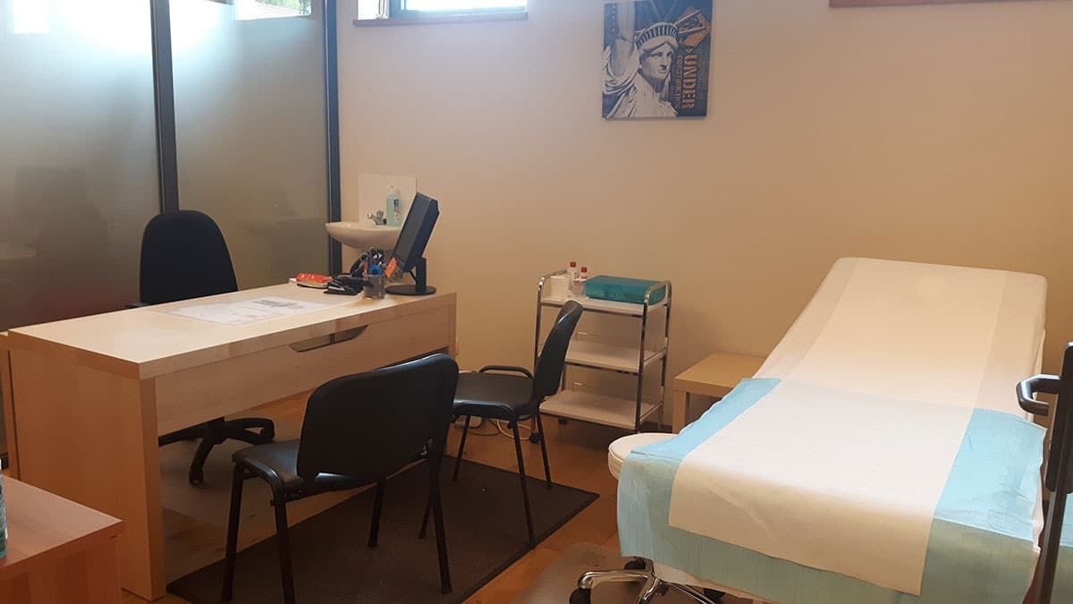 Salle d’examen médical avec deux tables, des chaises et une table d’examen recouverte d’un drap blanc, près d’une fenêtre.