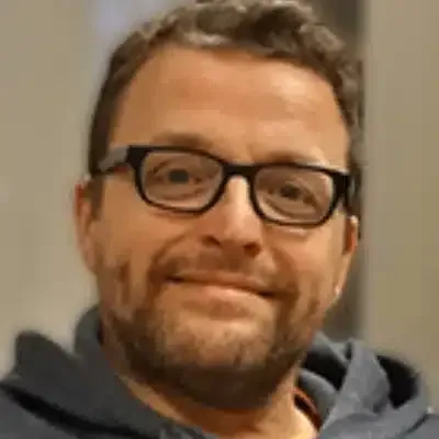 Un homme souriant portant des lunettes et un sweat à capuche gris, sur fond intérieur flou