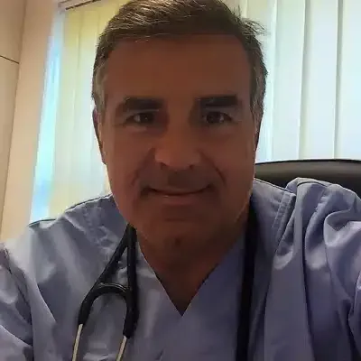 Un professionnel de santé en blouse bleue, avec un stéthoscope, sourit dans une pièce à la lumière tamisée, près de rideaux.