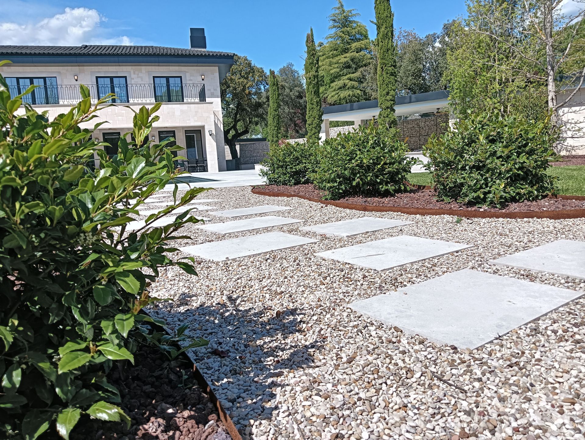 Patio de una casa moderna con grava, losas para caminar, arbustos y árboles altos bajo un cielo azul