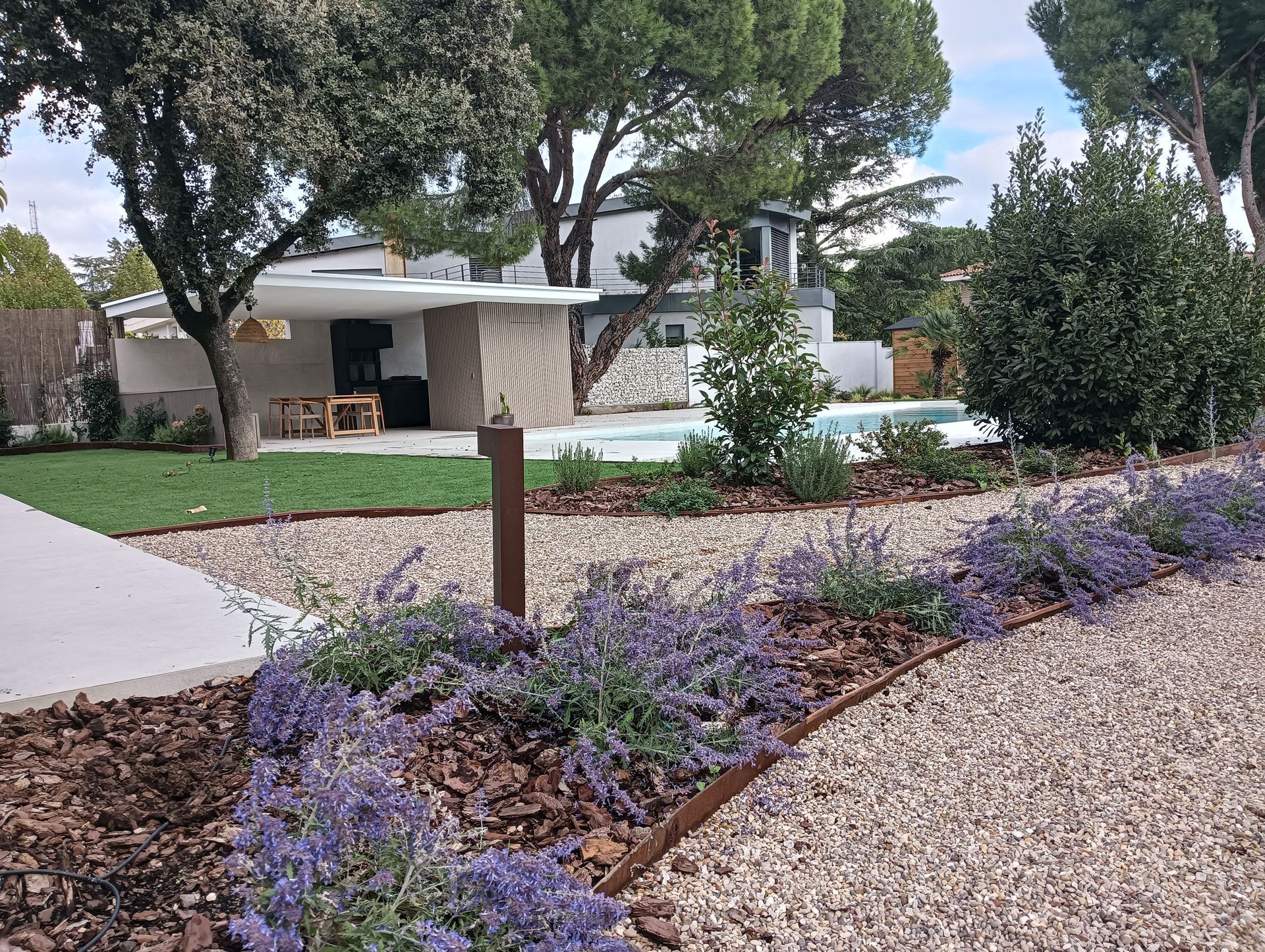 Patio ajardinado con flores moradas, senderos de grava, árboles y el patio de una casa moderna al fondo