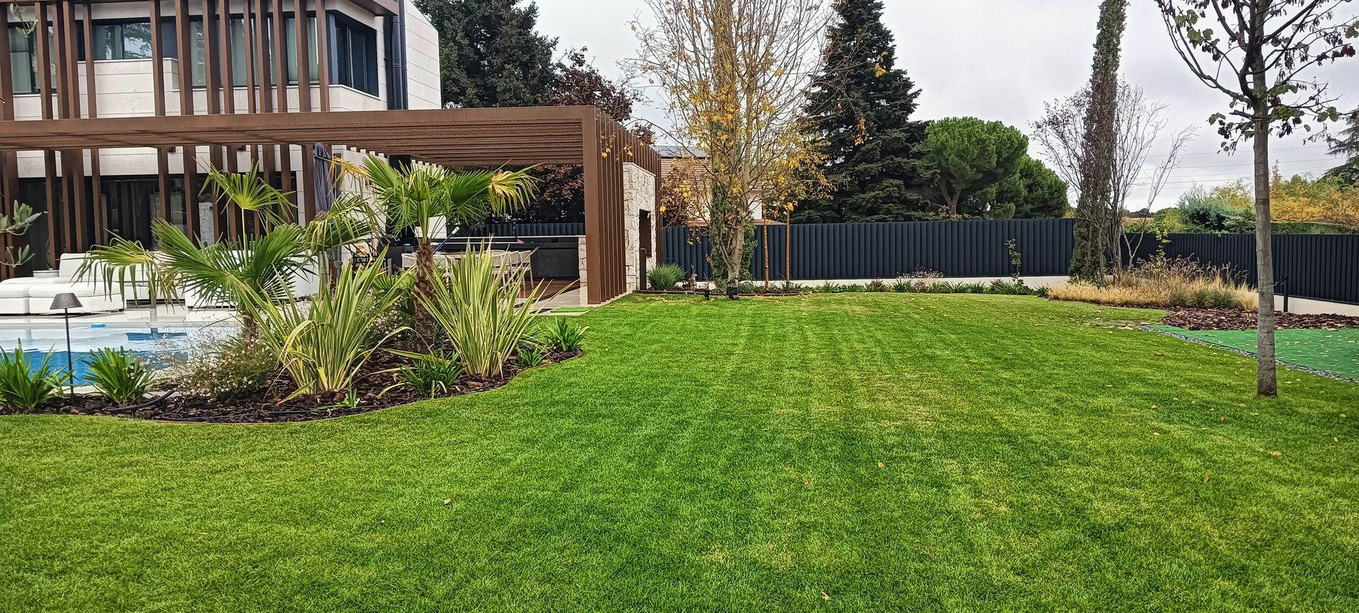 Jardín trasero moderno con césped verde, terraza de madera y piscina junto a un jardín cercado.