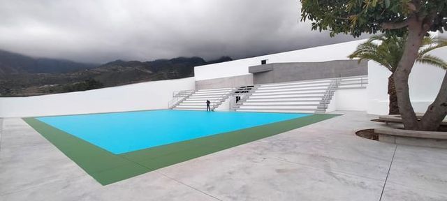 Una piscina moderna con agua azul y borde verde, junto a una pared blanca y un árbol, con montañas nubladas al fondo.