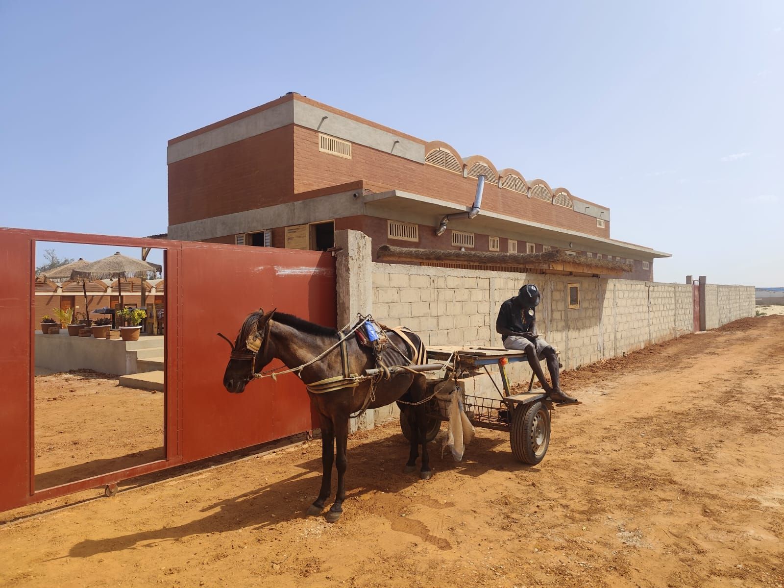 Carro tirado por caballos frente a una puerta roja junto a un edificio de ladrillos en un camino de tierra.