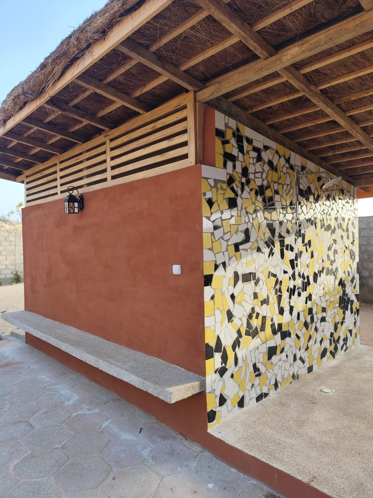 Baño exterior o caseta de servicio con estuco rojo, pared de mosaico y techo inclinado bajo un cielo brillante.