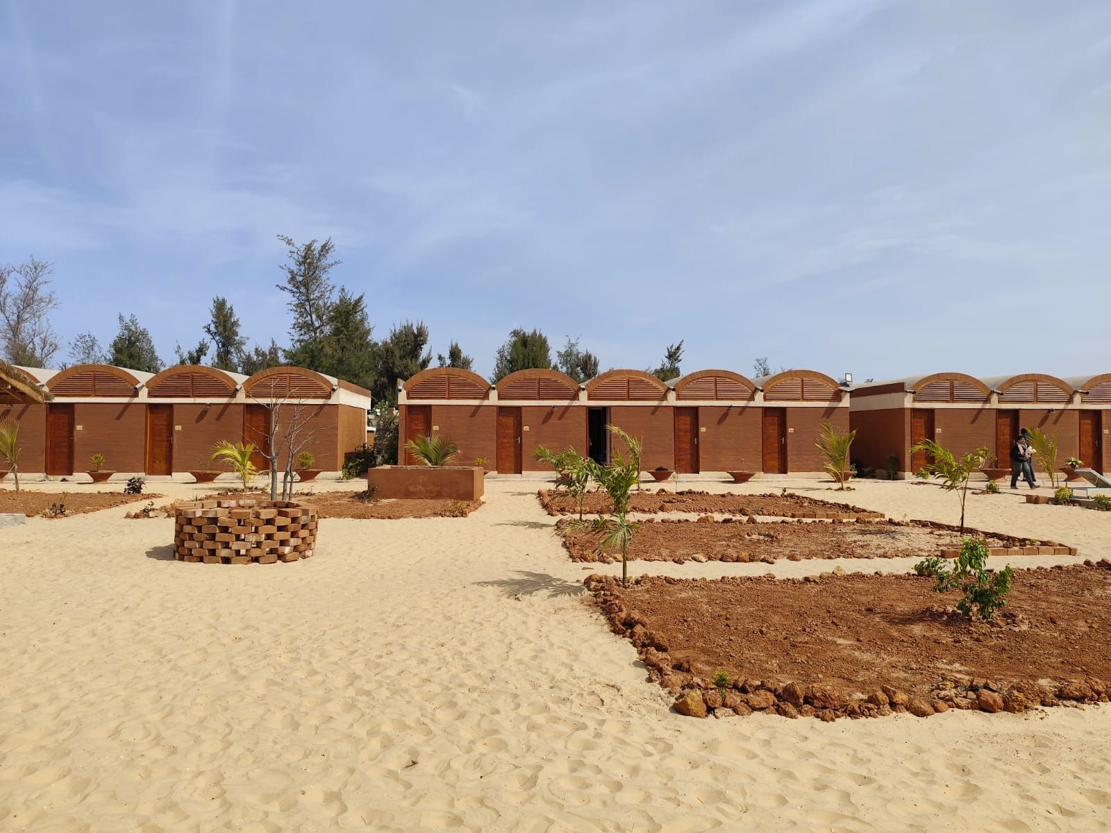 Campamento en el desierto arenoso con hileras de tiendas marrones y pequeños arbustos plantados bajo un cielo nublado.
