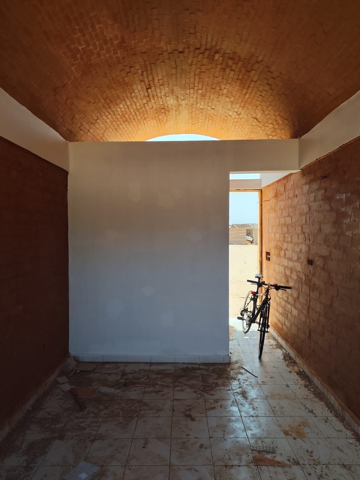 Pasillo estrecho con paredes de ladrillo, tabique blanco, suelo de baldosas y una bicicleta cerca de la luminosa puerta.