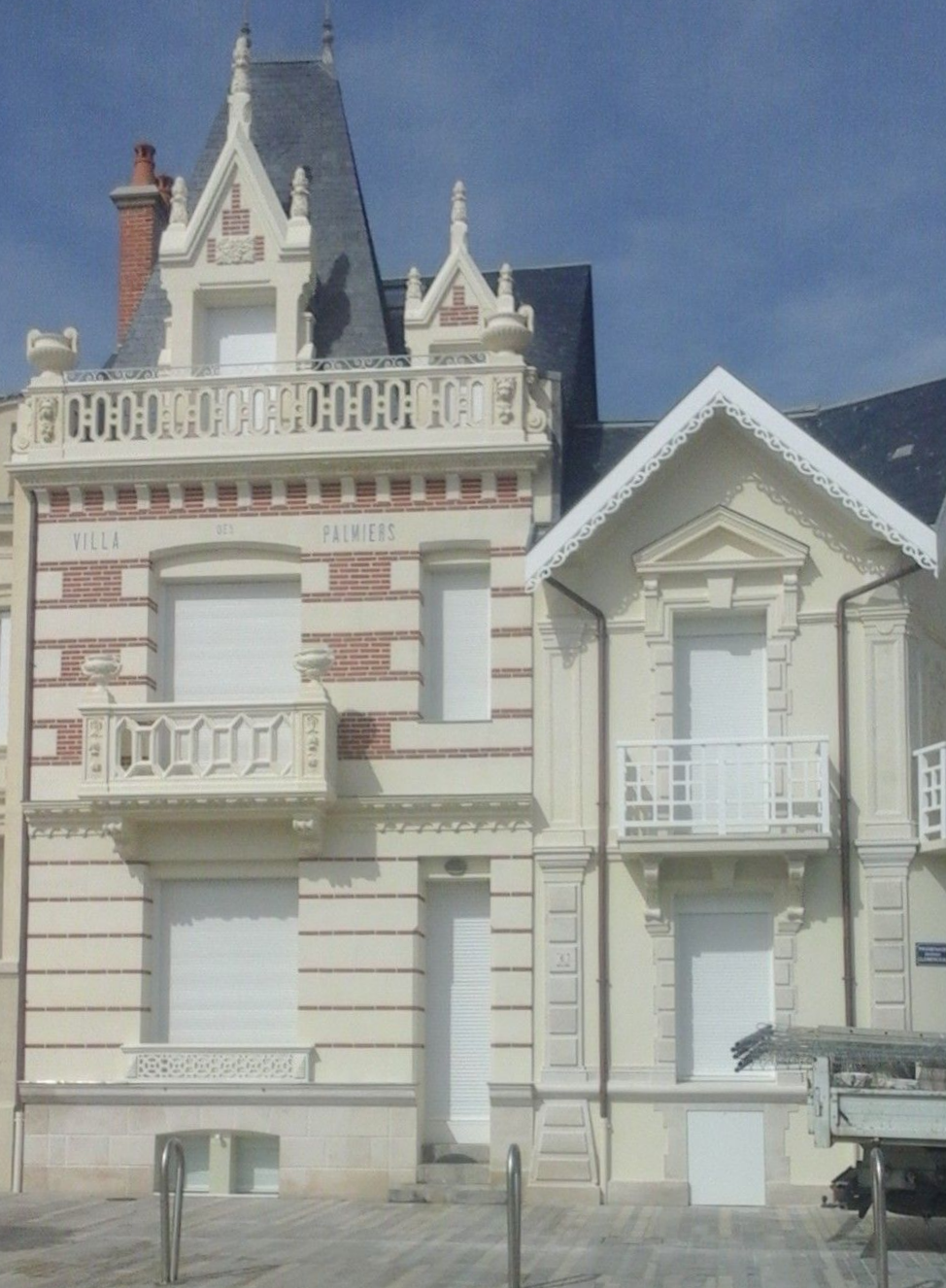 Deux bâtiments de deux étages, l'un en briques rouges avec des ornements blancs et un balcon, l'autre à façade blanche, tous deux sous un ciel bleu.
