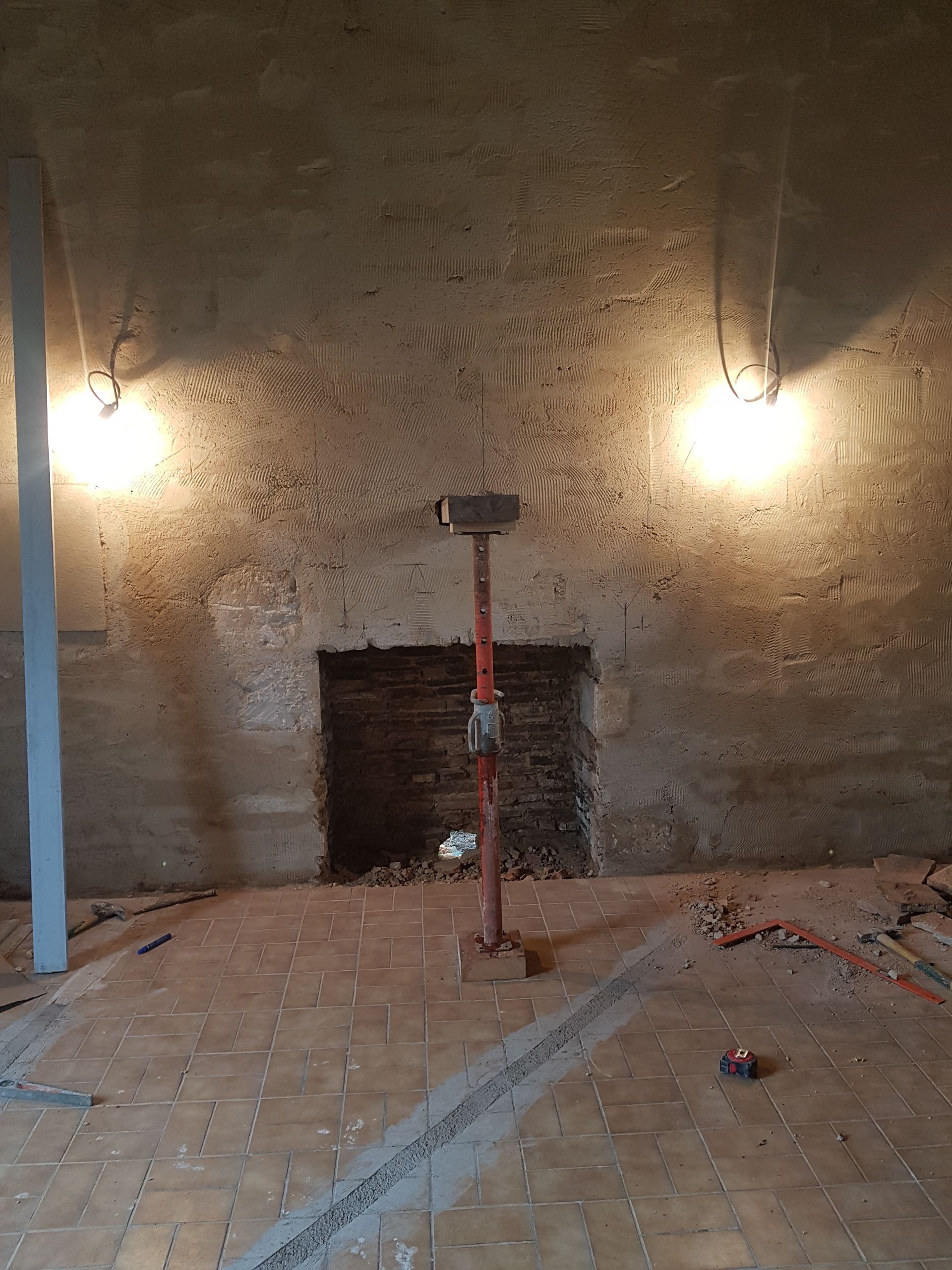 Pièce en rénovation avec briques apparentes, deux luminaires, une poutre de soutien et des débris de construction.
