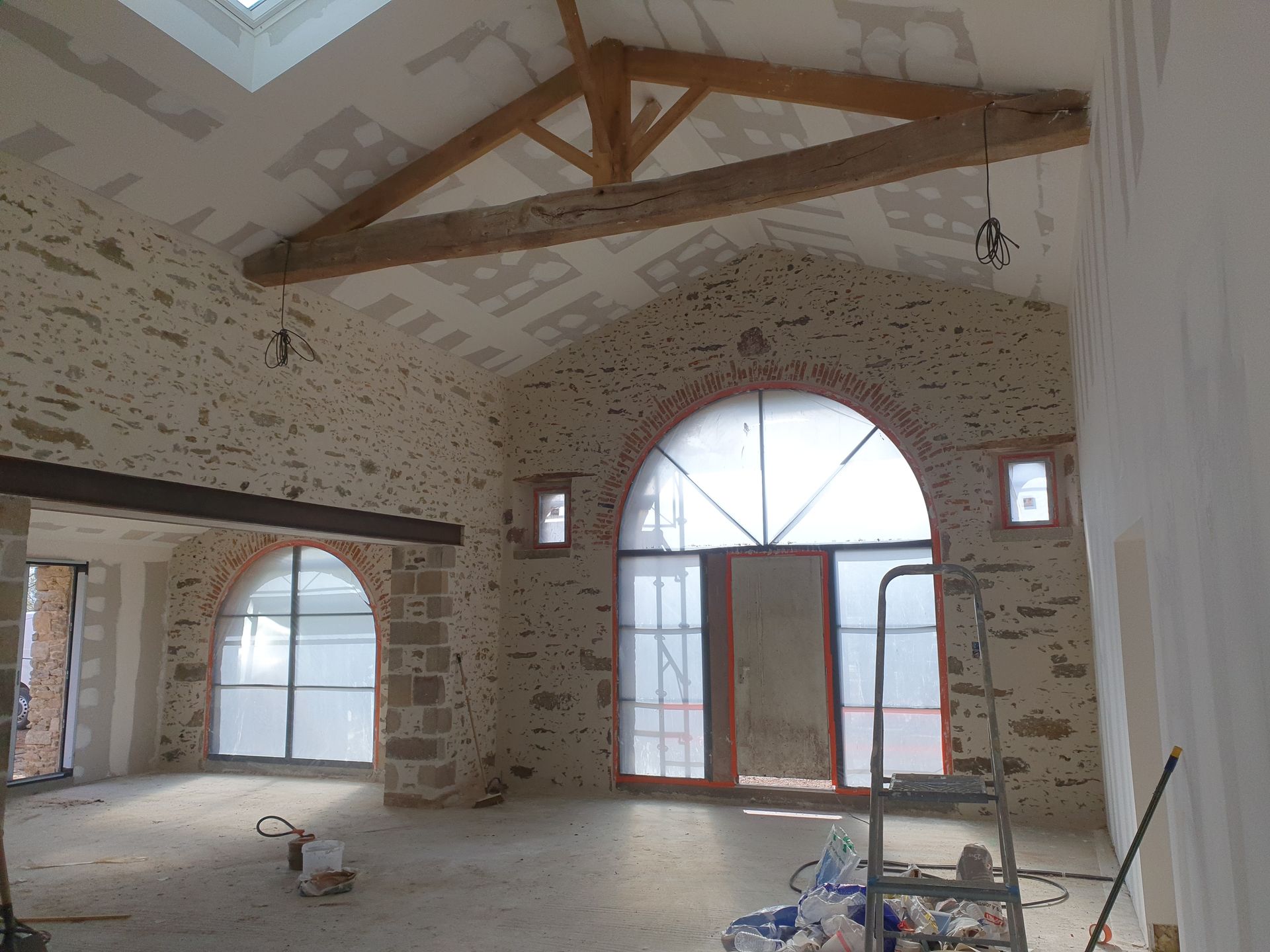 Intérieur d'une pièce aux murs de pierre avec fenêtres cintrées, poutres en bois et puits de lumière. Travaux en cours.