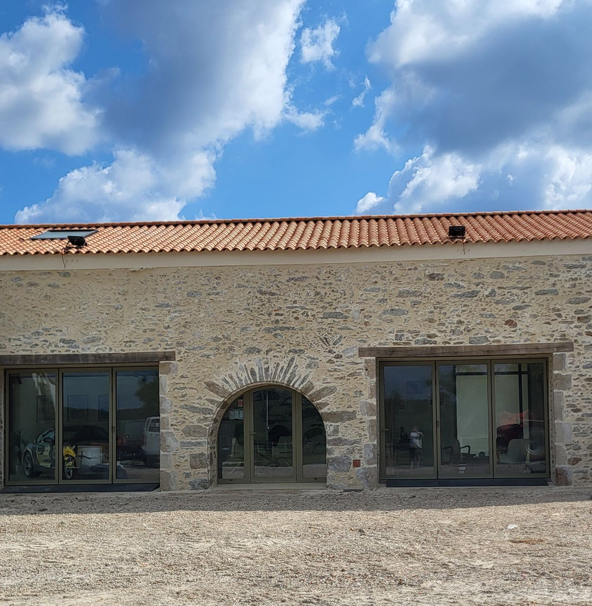Bâtiment en pierre avec une fenêtre cintrée et deux grandes fenêtres rectangulaires, toit de tuiles rouges, ciel bleu.