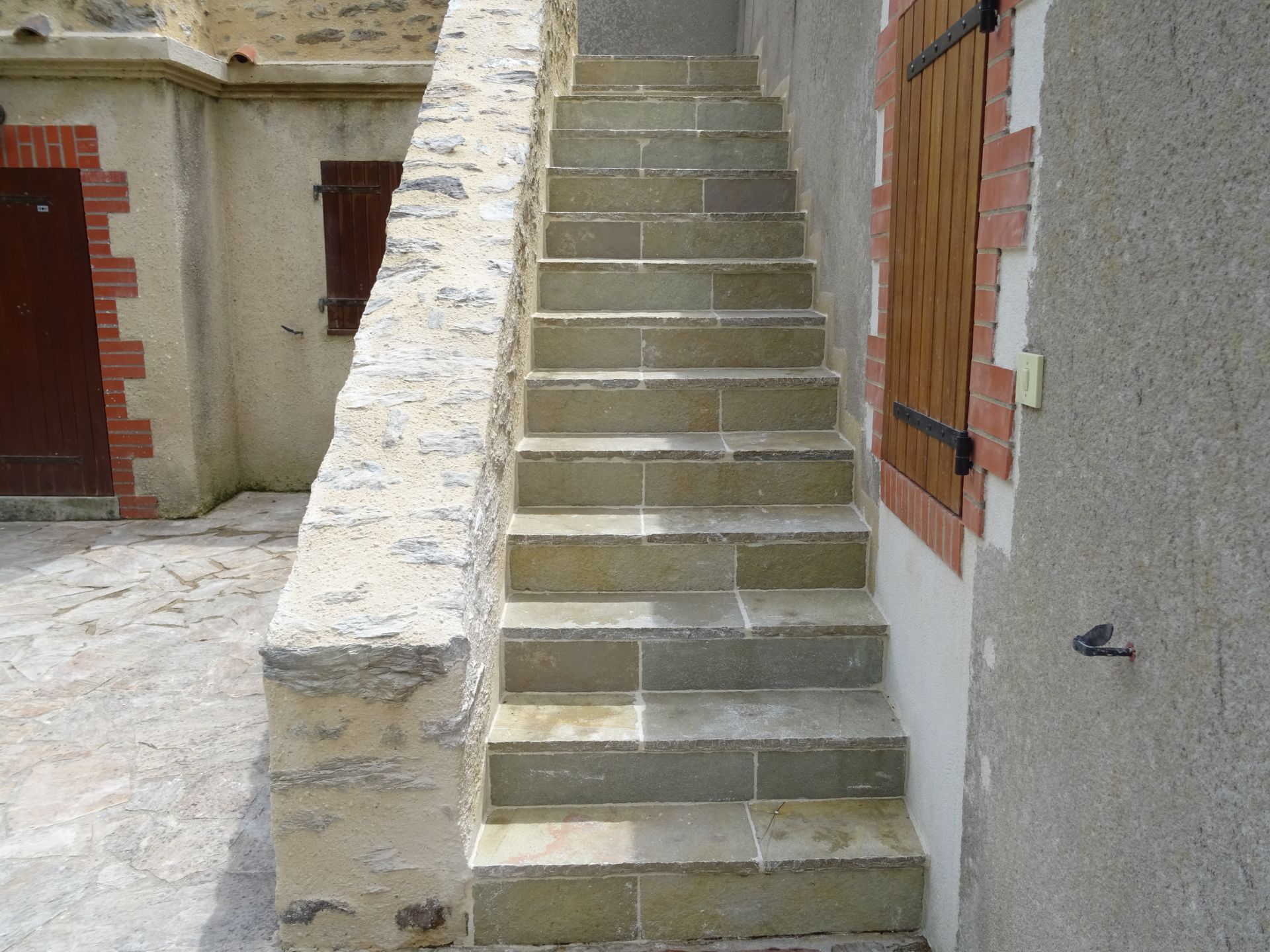Escalier extérieur en pierre avec une rampe en pierre et façade du bâtiment avec une porte et des volets bruns.