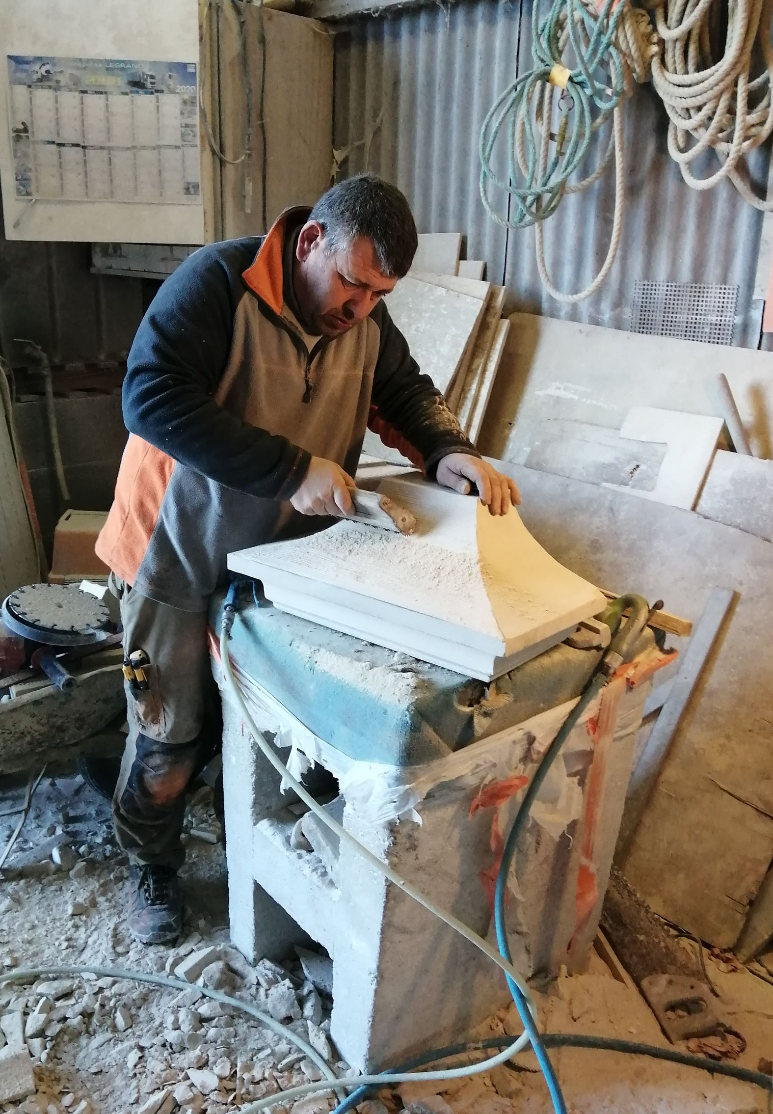 Un homme sculpte la pierre, vêtu d'orange et de noir, à l'intérieur, utilisant un outil sur une structure en pierre blanche.