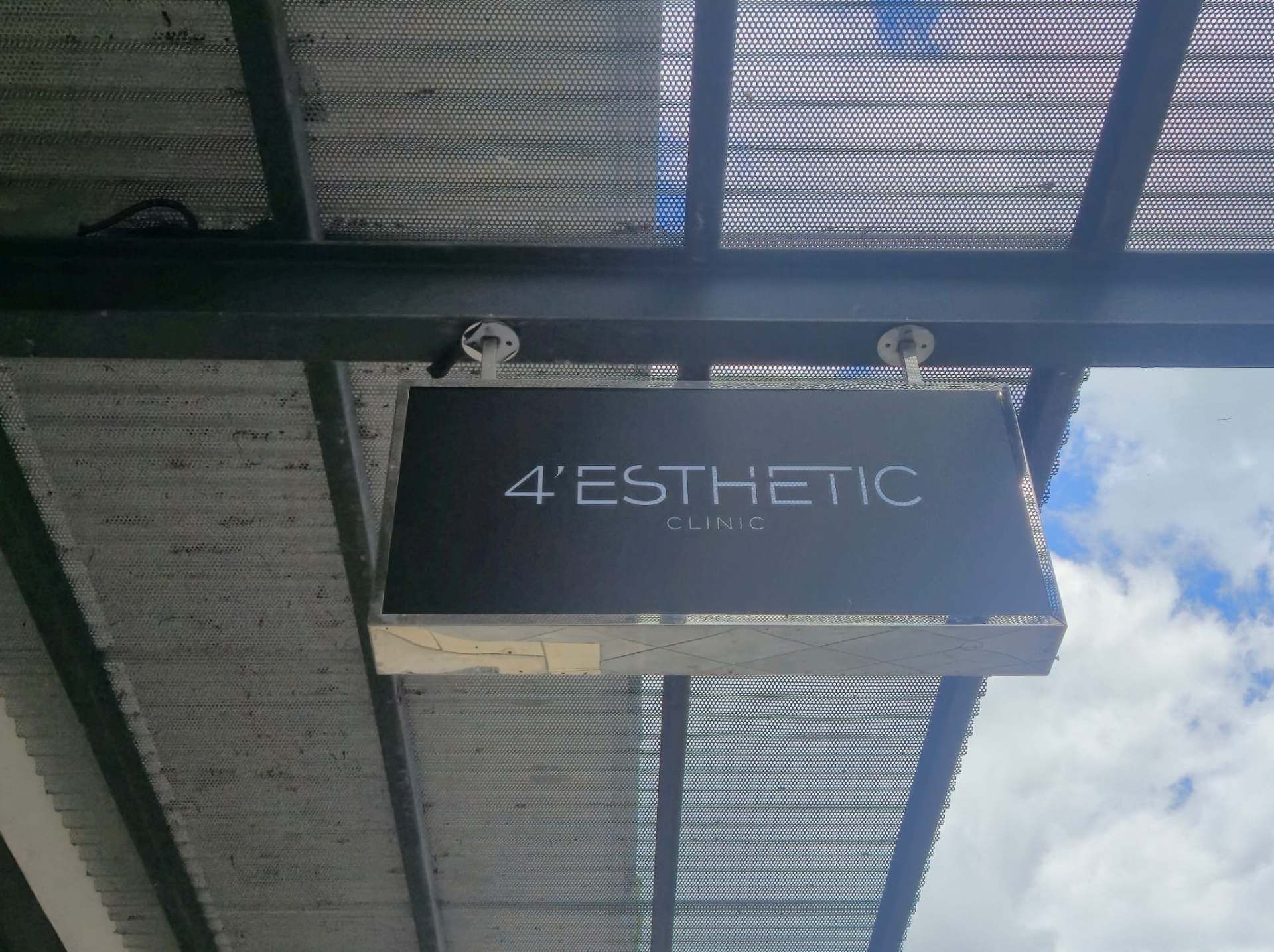 Schwarzes Hängeschild mit der Aufschrift „4'ESTHETIC“ unter einem Metalldach vor dem Himmel