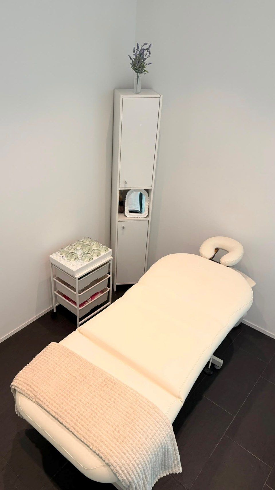 Leerer Massagebehandlungsraum mit beiger Liege, weissem Schrank und gestapelten Handtüchern in einer sauberen Ecke.