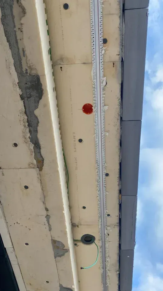 Unterseite der Dachtraufe eines Gebäudes mit freiliegender Isolierung, einem roten Sicherungskasten und Leitungen vor einem bewölkten Himmel.