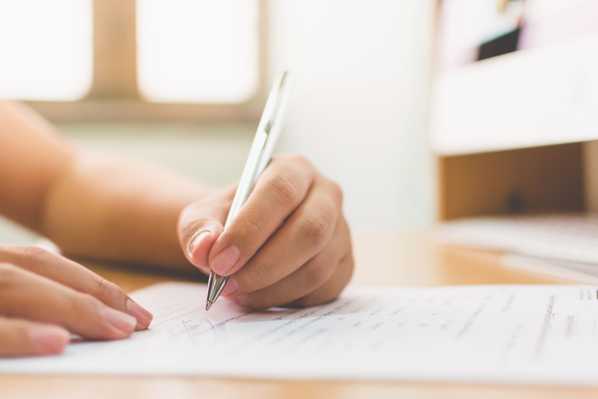 Die Hand einer Person schreibt mit einem Stift auf Papier auf einem Schreibtisch, im Hintergrund ein Computer.