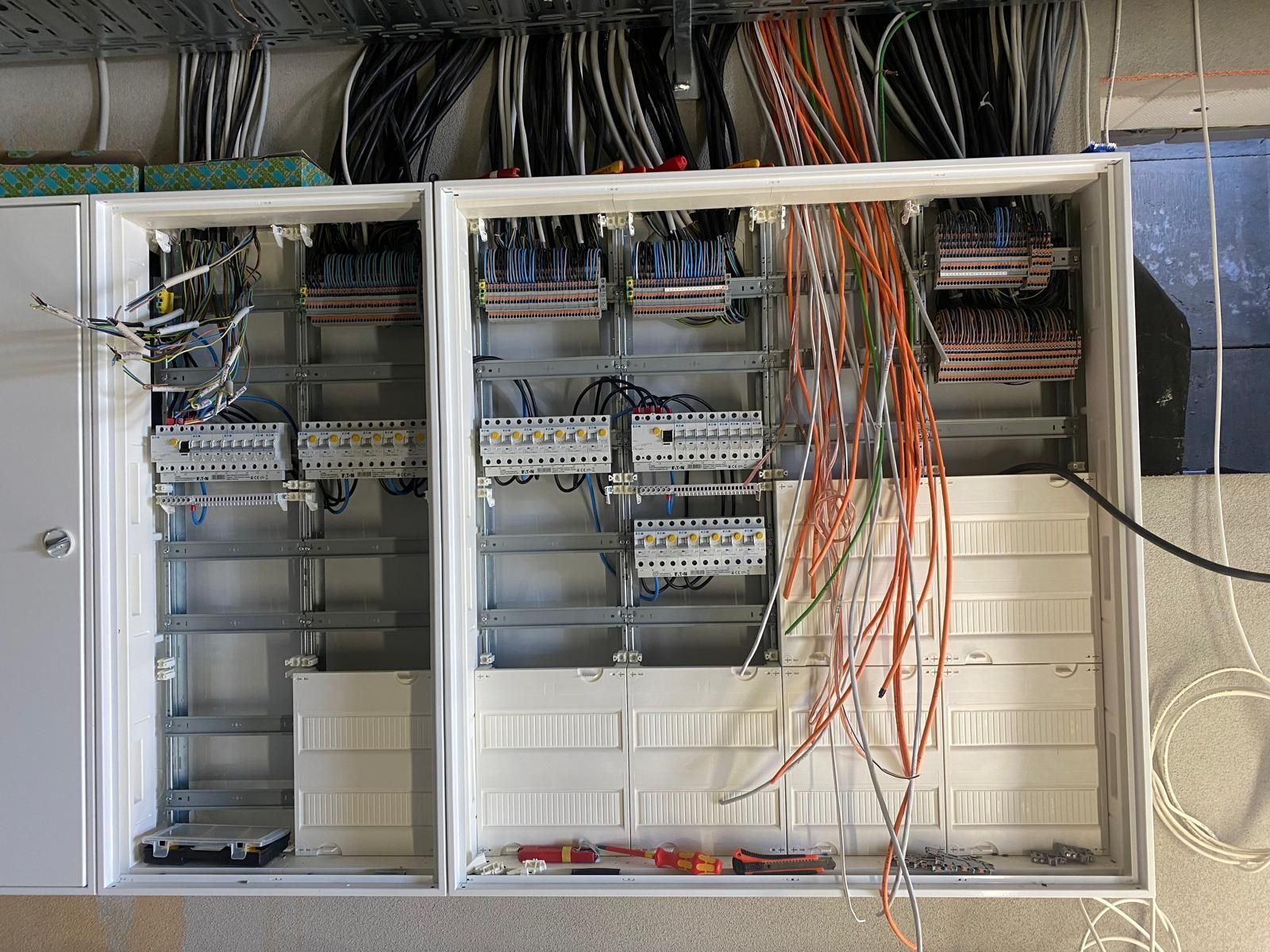 Schalttafel mit freiliegenden Kabeln in einem grauen Schrank, die Kabel sind orange, schwarz und weiß.