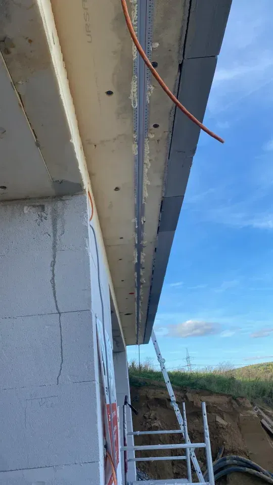 Baustelle: Eine Dachkante mit dunkelgrauer Eindeckung, hellgrauem Beton und einer teilweise errichteten Ziegelwand; eine Leiter ist sichtbar.