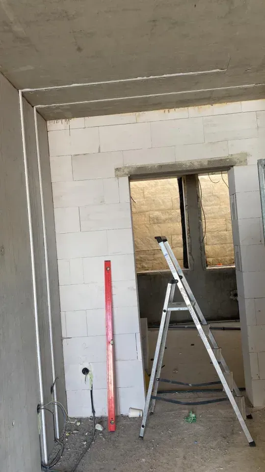 Baustelle: Leiter im Türrahmen, waagerechte Neigung, freiliegende Betonwand, unfertiger Raum.