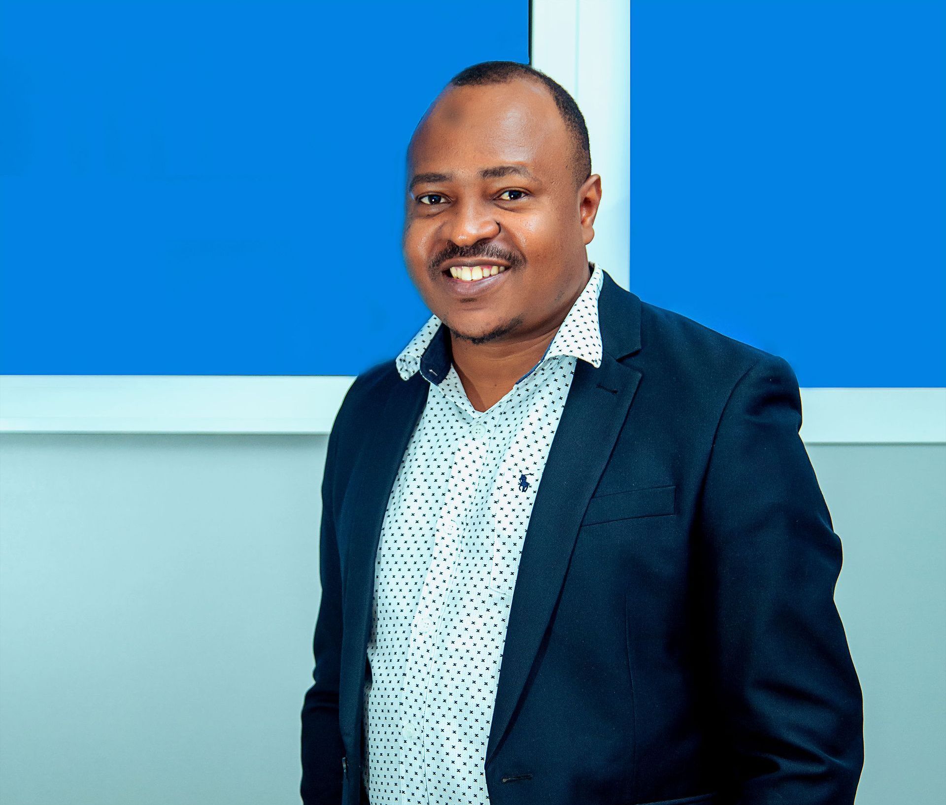 A man in a suit and polka dot shirt is smiling in front of a blue wall.