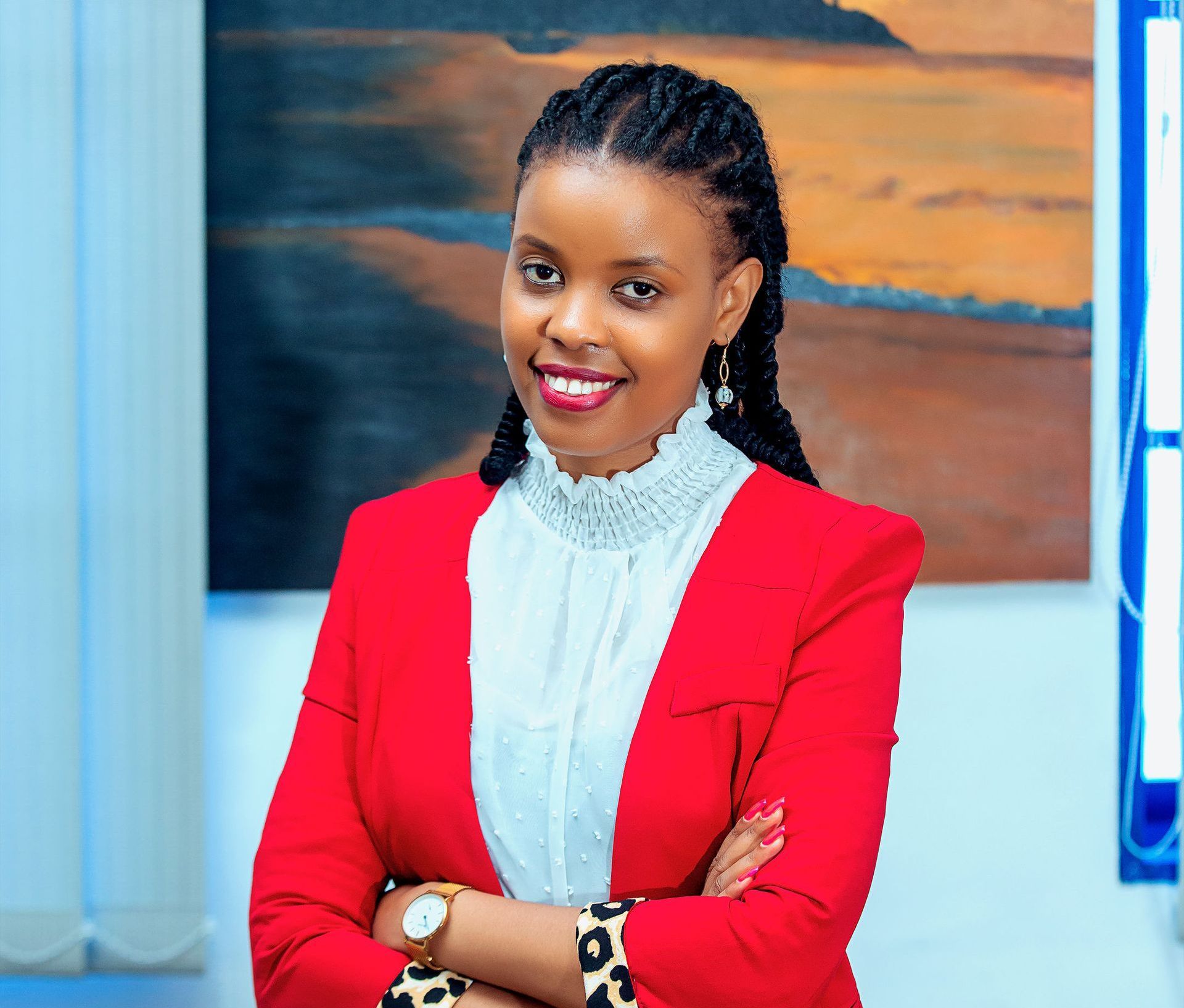 A woman in a red jacket and white shirt is standing with her arms crossed in front of a painting.