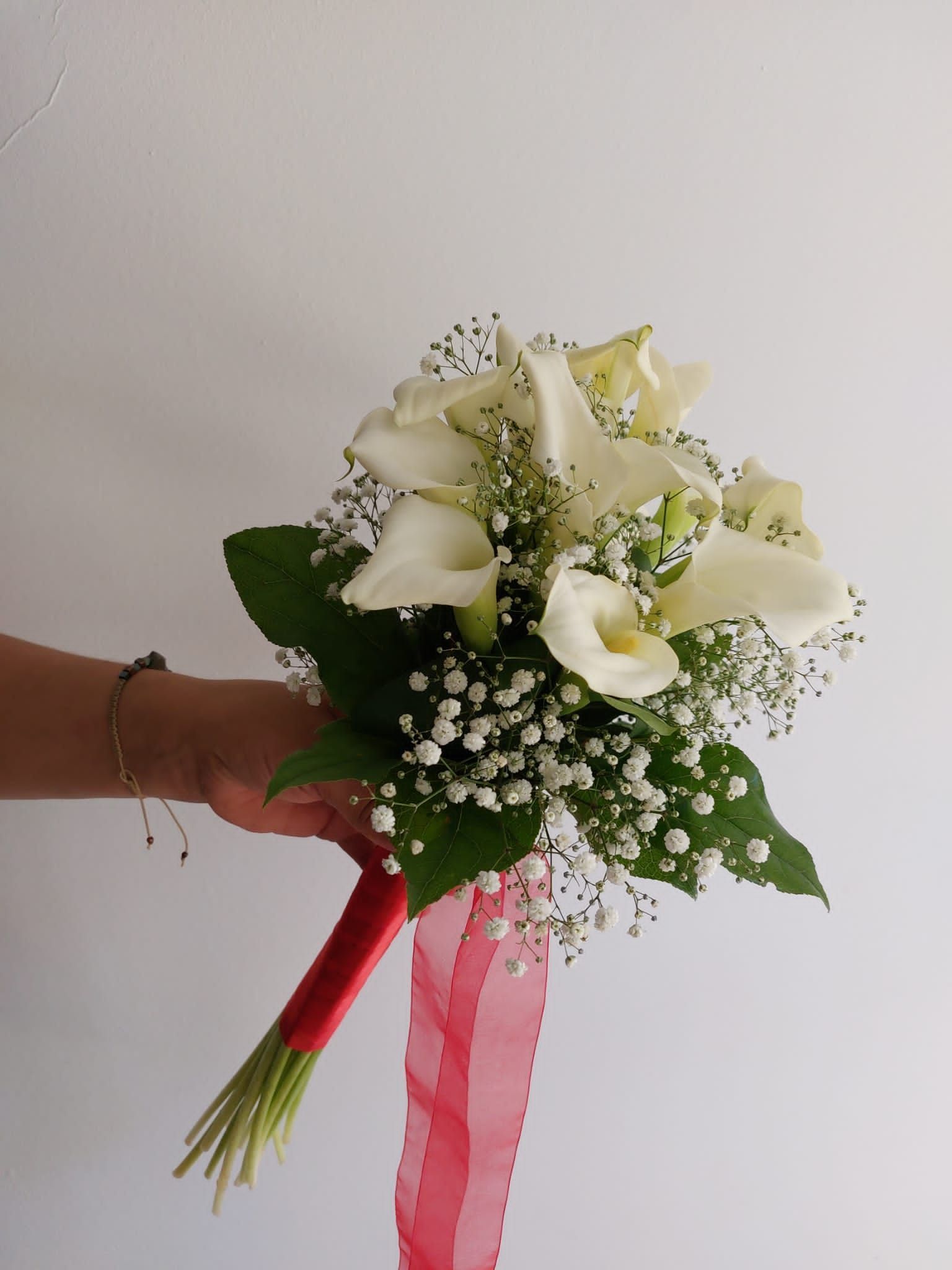 Una mano sosteniendo un ramo de novia de calas blancas y flores de bebé, con una cinta roja y un fondo blanco.