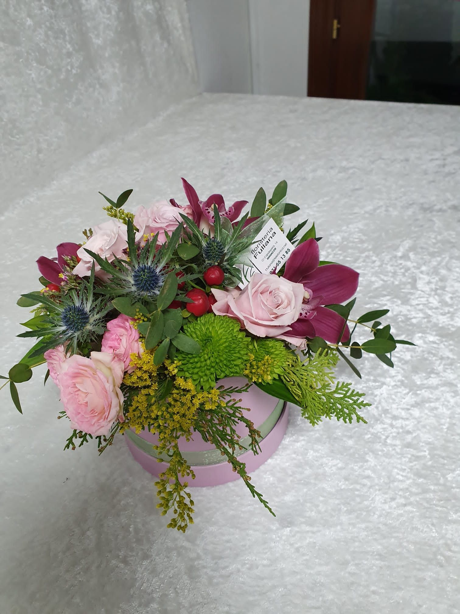 Un arreglo floral en una caja rosa incluye rosas rosadas, flores moradas y follaje. Se coloca sobre una superficie blanca texturizada.