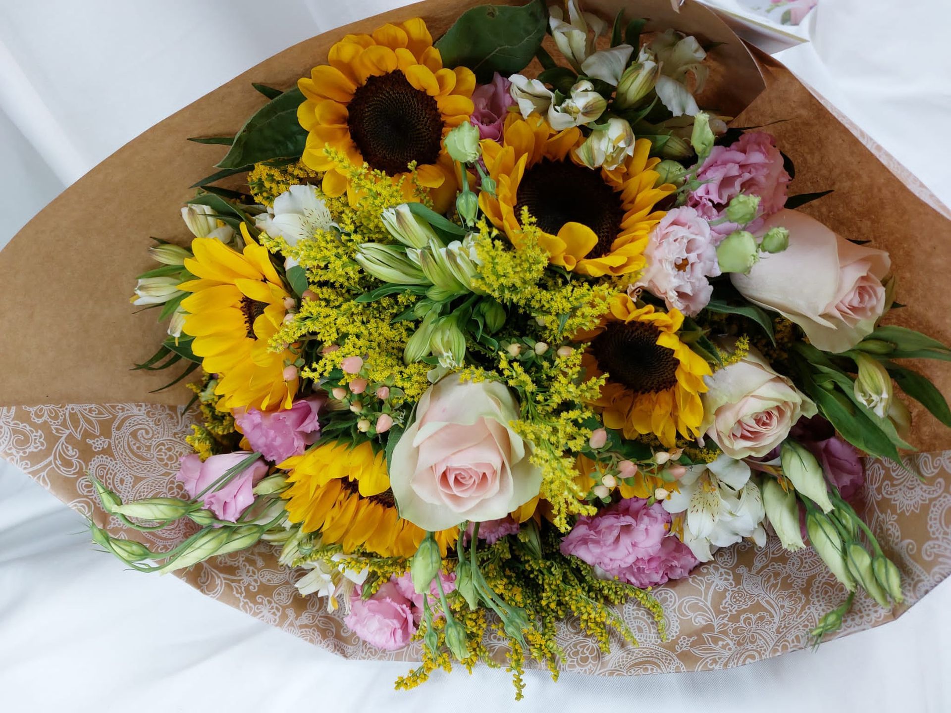 Un ramo de girasoles amarillos brillantes, rosas rosadas y flores blancas envuelto en papel marrón.
